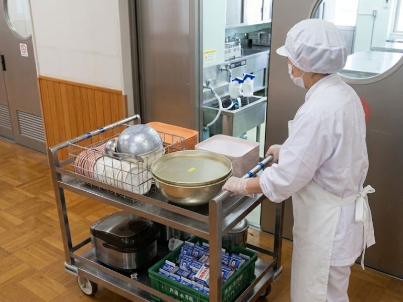 白い調理服とエプロン、帽子、マスクを身につけたスタッフが、食器や牛乳が載った配膳台を慎重に押して廊下を運んでいる様子。